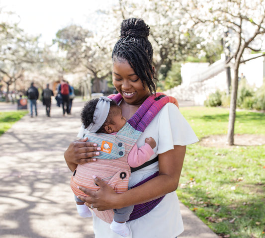 A mother holding her baby in a Signature baby carrier while being in a park.