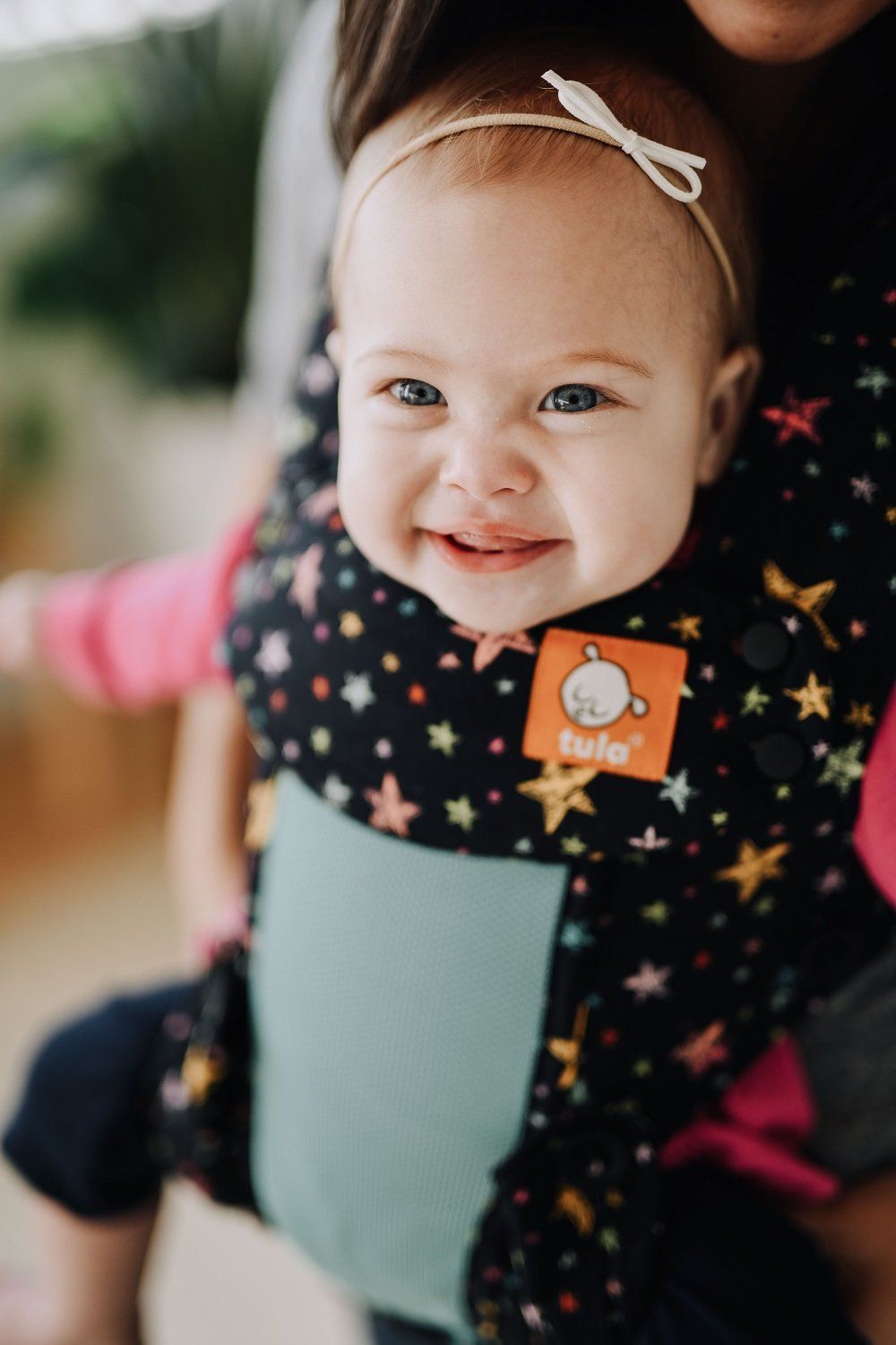 A baby smiling in the camera while sitting in the Explore Baby Carrier Coast Rainbow Stars.