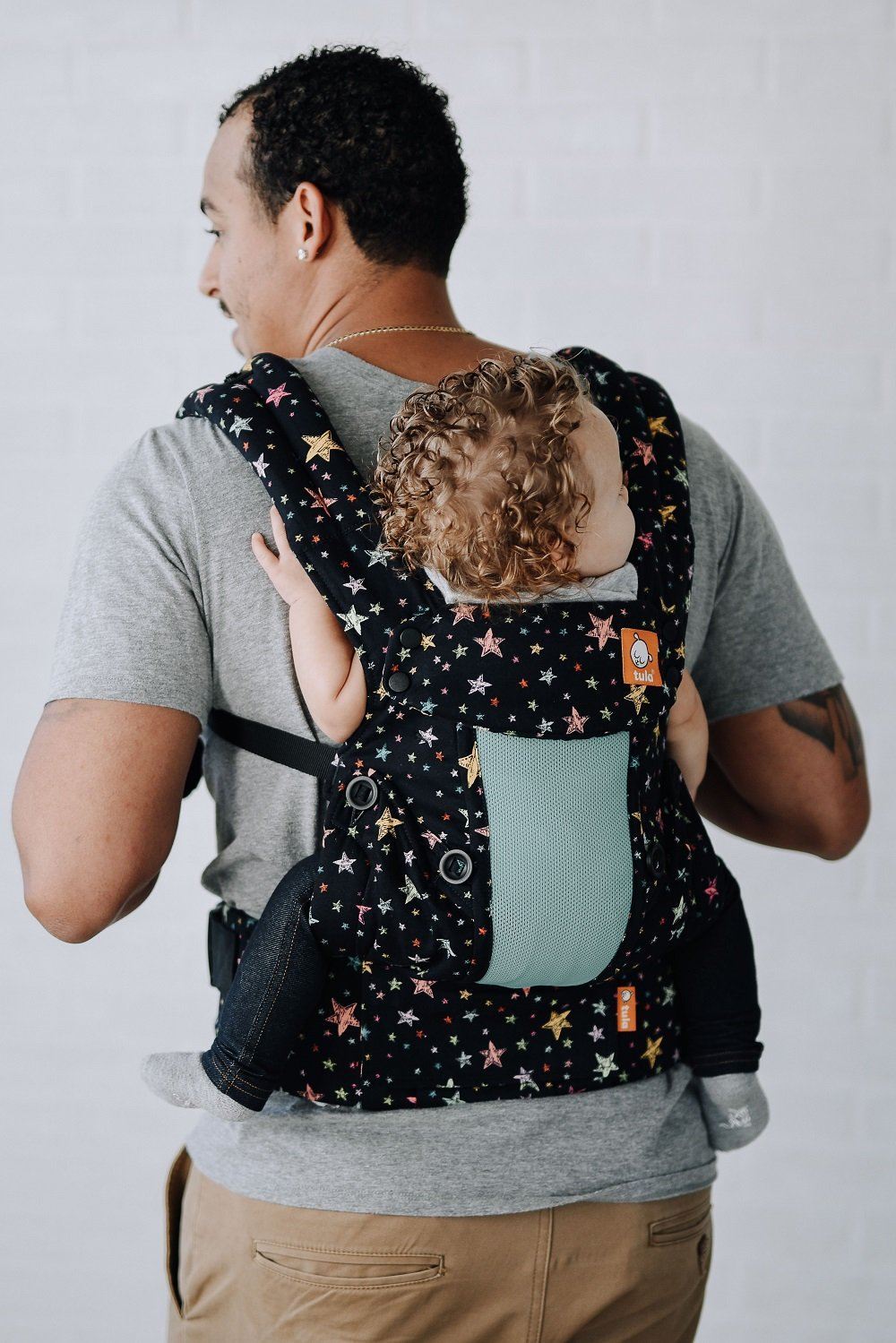 A baby sitting in the Explore Baby Carrier Coast Rainbow Stars in back-carry position.