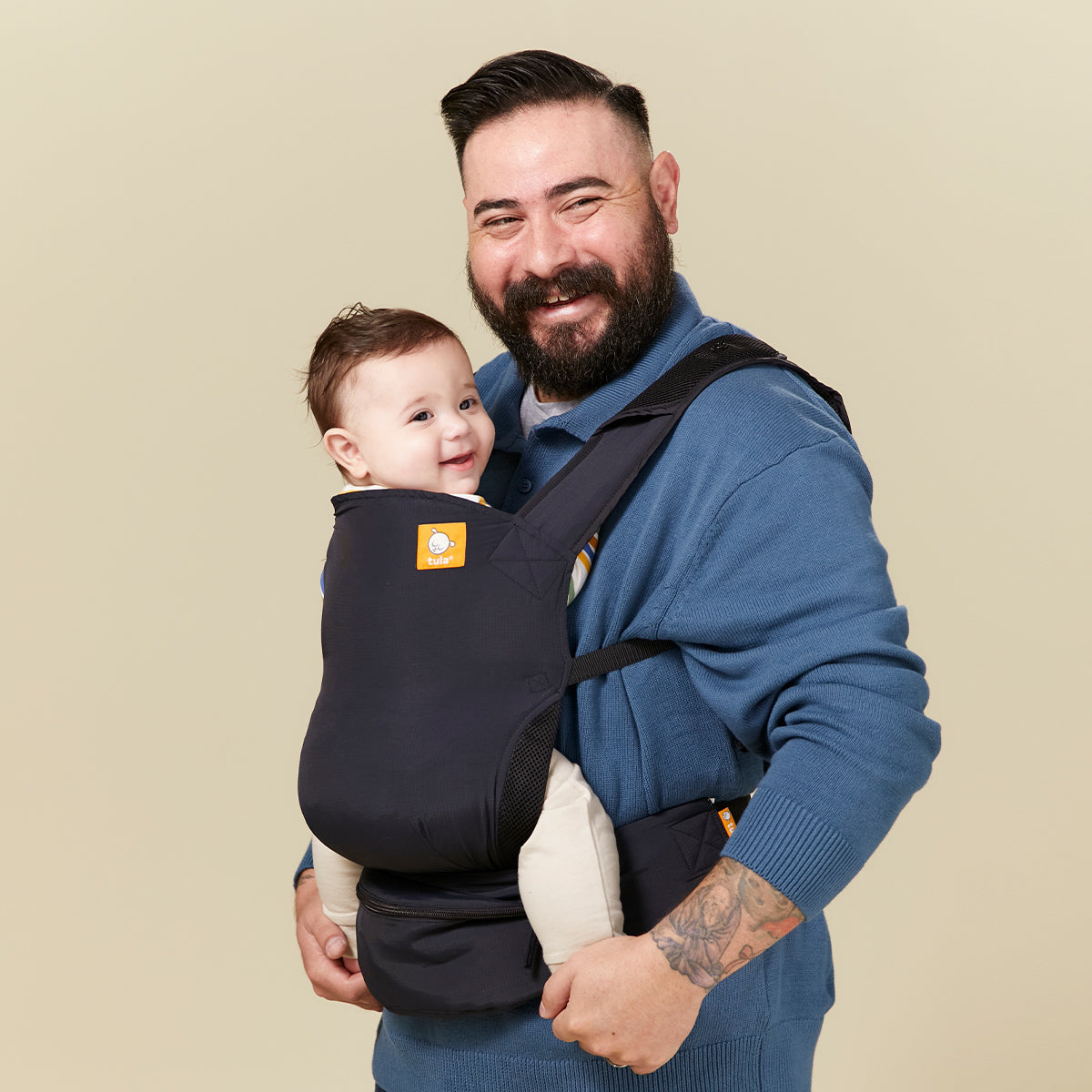 Un papá sonriente lleva a su hijo feliz en la mochila porta bebé Tula Lite Negro