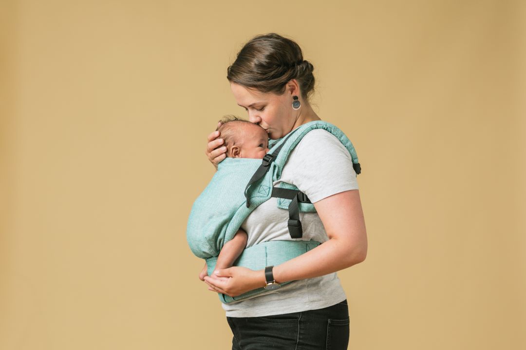 Una mamá besa a su bebé recién nacido en un portabebés ergonómico desde el nacimiento