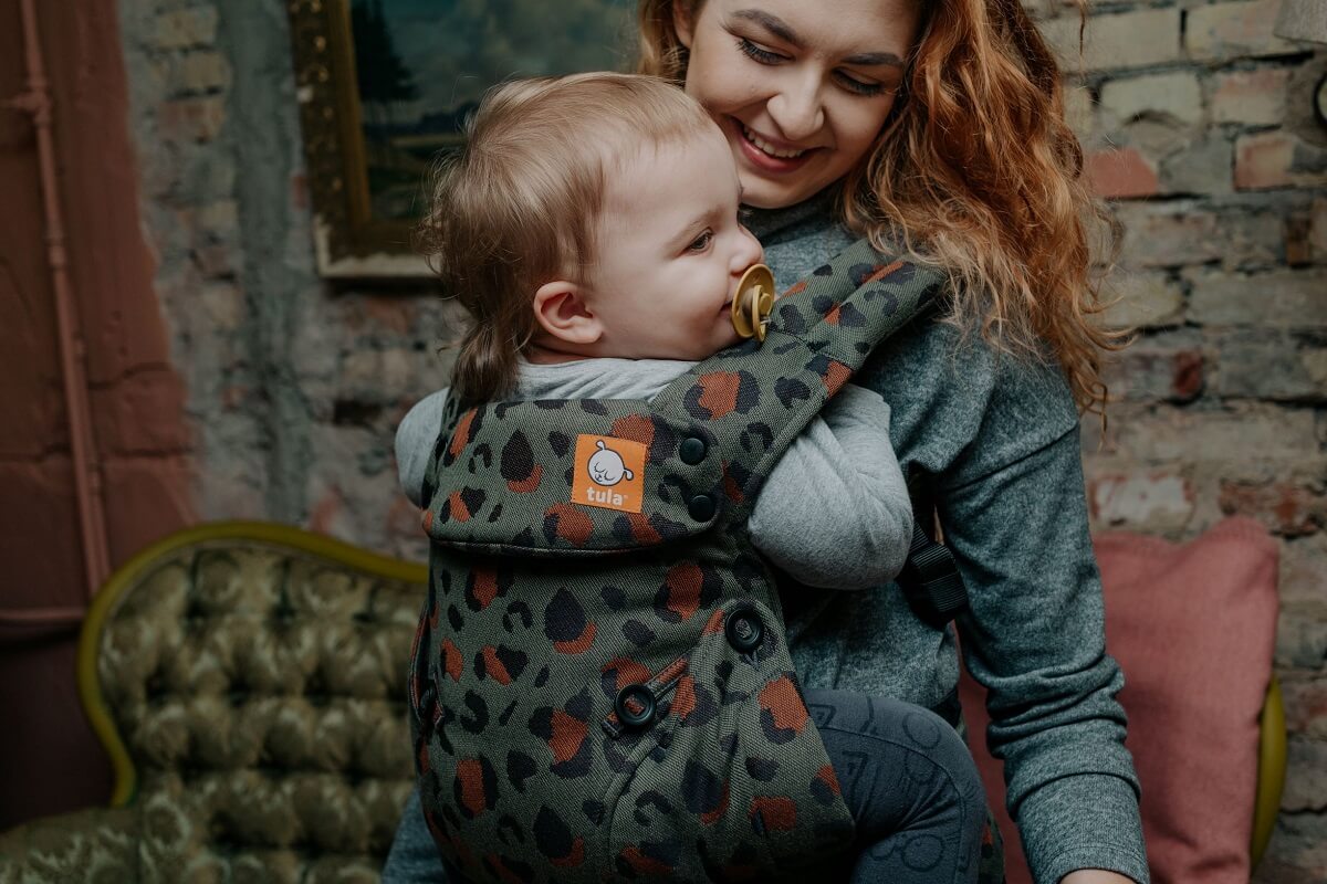Une mère souriante porte son enfant dans le porte-bébé ergonomique Signature Explore  Olive Leopard  en position de portage avant.