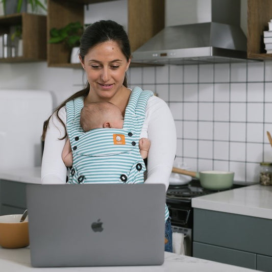 Uma mãe carrega o seu bebé adormecido num Porta-bebés enquanto trabalha no seu Mac Book.